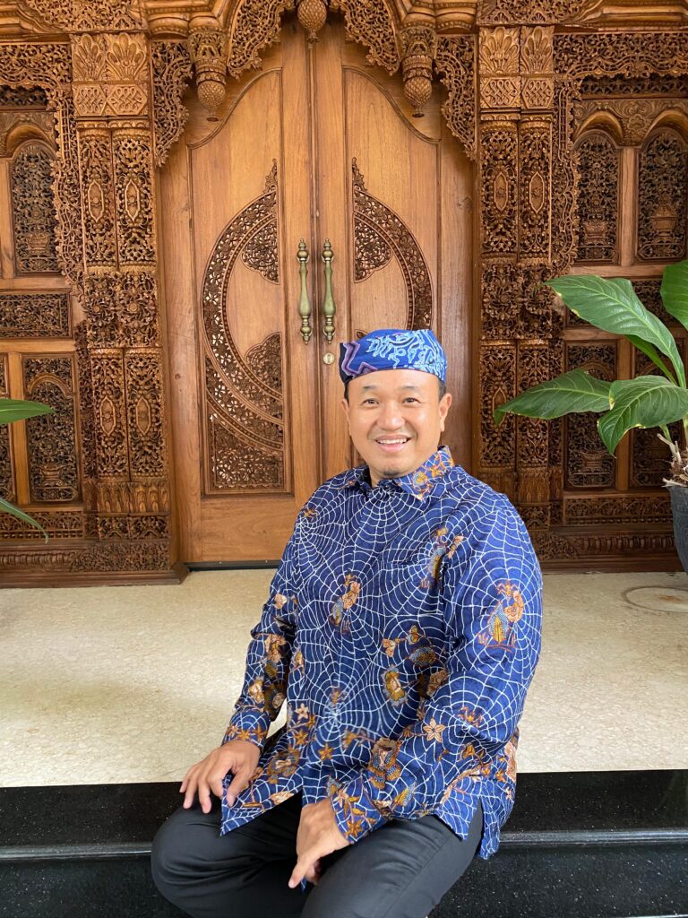 Whatsapp image 20260609 at 13 46 47 catrawarta Smiling man in a blue batik shirt and matching headscarf sits on steps in front of an intricately carved wooden door with floral motifs behind him